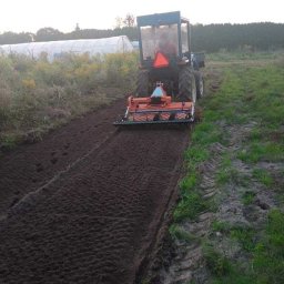 AGRO-PARK - Traktor z glebogryzarką spulchnia ziemię na polu, przygotowując ją pod uprawę. Widoczne świeżo obrobione pasy ziemi i ślady opon. W tle szklarnia.