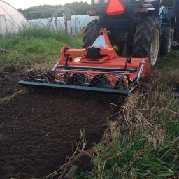 AGRO-PARK - Pomarańczowy traktor z glebogryzarką w trakcie pracy na polu, przygotowujący glebę pod uprawę. W tle widoczny tunel foliowy.