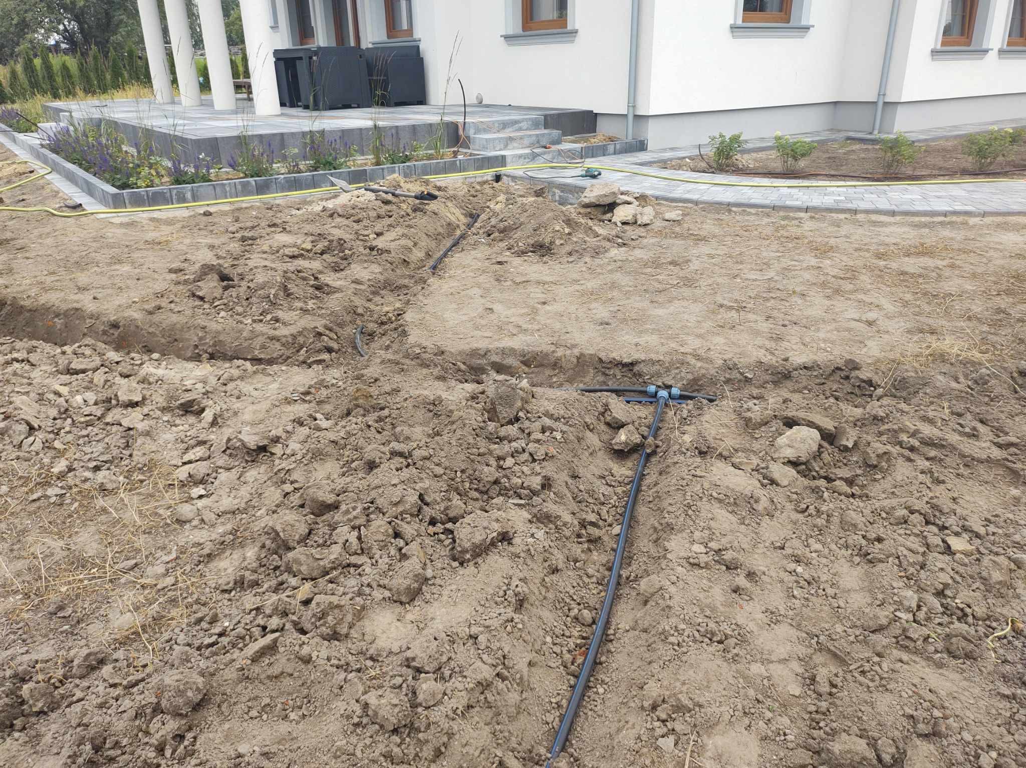 Wykopana ziemia z widocznymi rurami instalacji nawadniającej, fragment domu z kostką brukową w tle. Etap prac przy zakładaniu systemu nawadniania ogrodu.