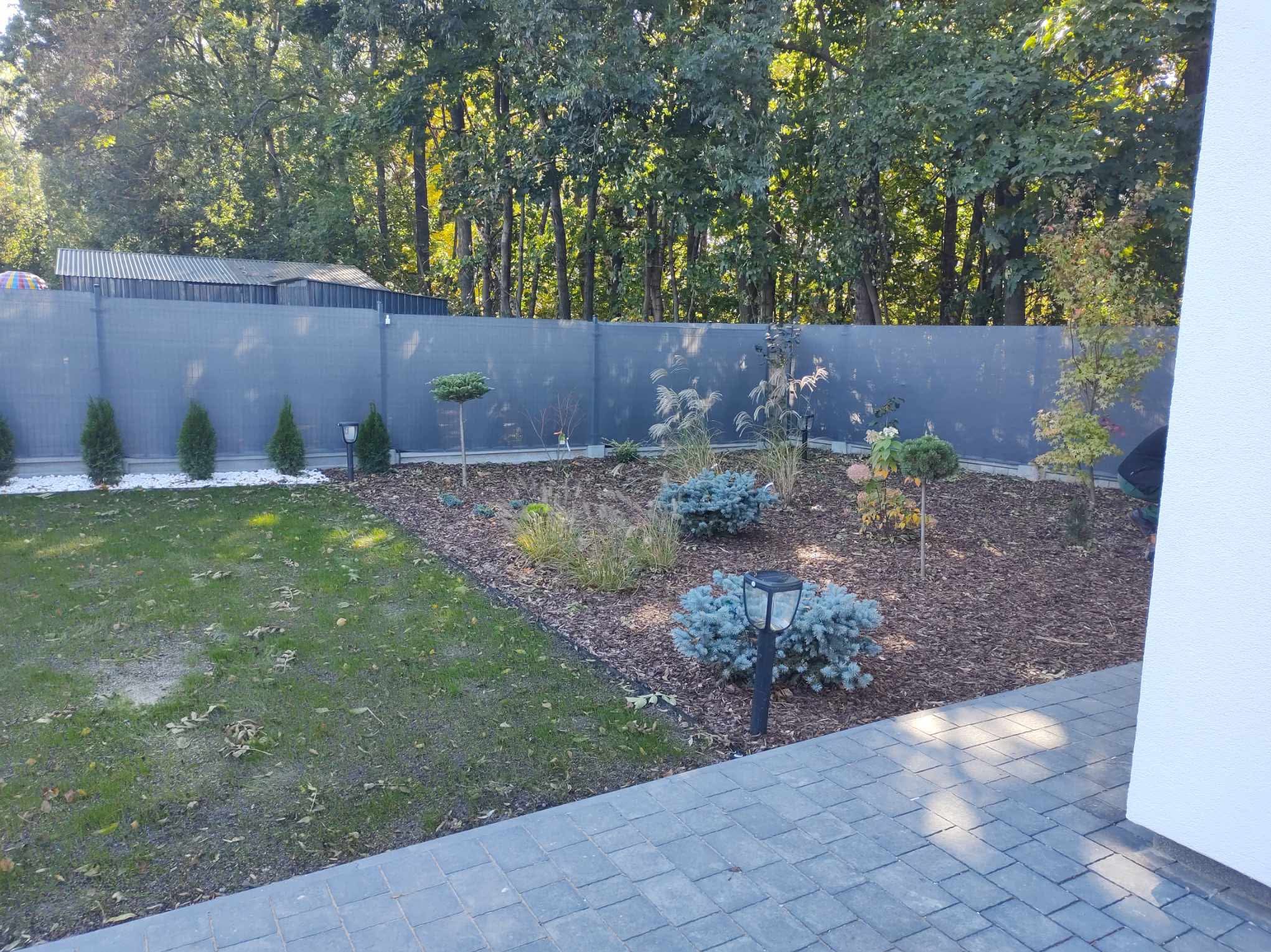 Uporządkowany ogród z ozdobnymi krzewami, drzewami i korą. Widoczny trawnik, szary płot i lampy solarne. Całość tworzy estetyczną przestrzeń.