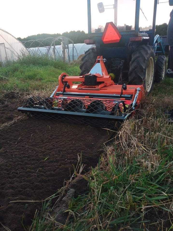 Pomarańczowy traktor z glebogryzarką w trakcie pracy na polu, przygotowujący glebę pod uprawę. W tle widoczny tunel foliowy.