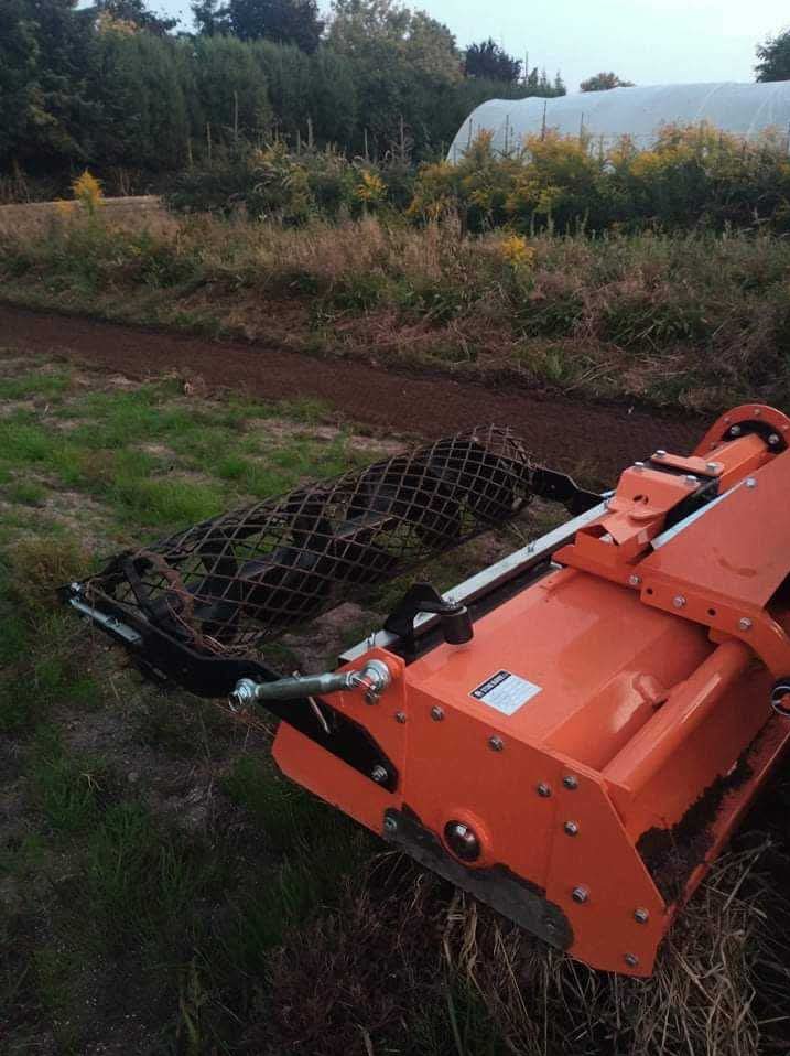 Pomarańczowy glebogryz z wałem strunowym na polu, przygotowujący ziemię pod uprawę. W tle widoczna roślinność i tunel foliowy.