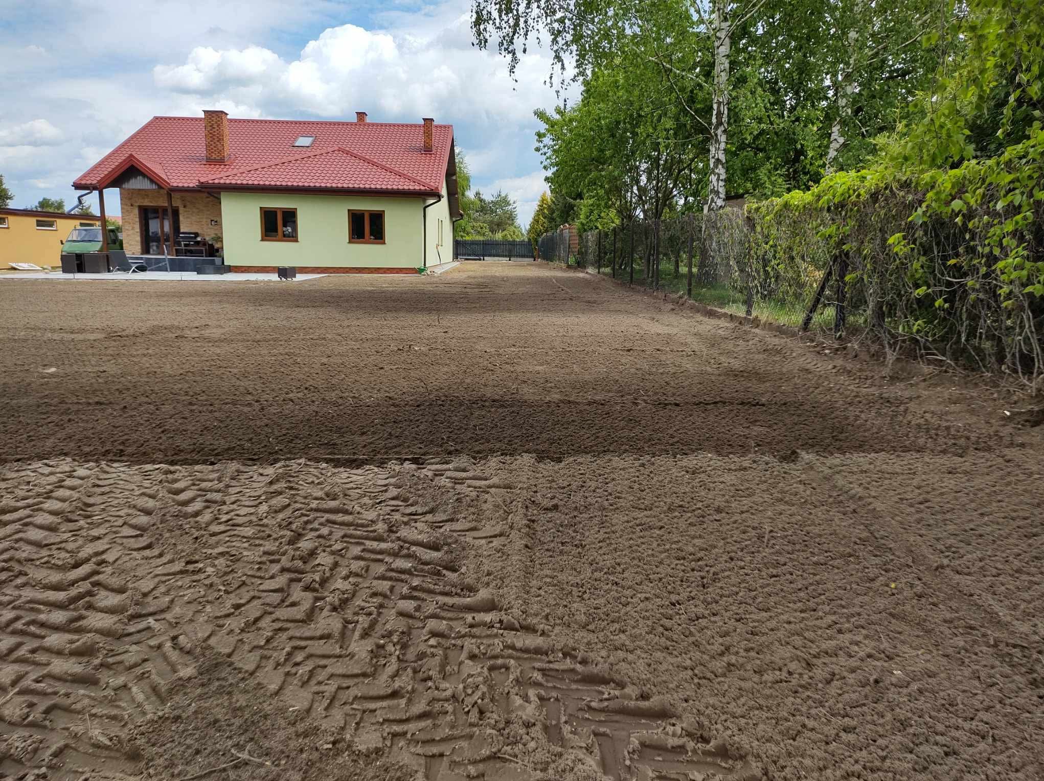 Przygotowany teren pod trawnik z widocznymi śladami opon, dom z czerwoną dachówką w tle. Grunt wyrównany, gotowy do siewu lub rozłożenia trawy z rolki.