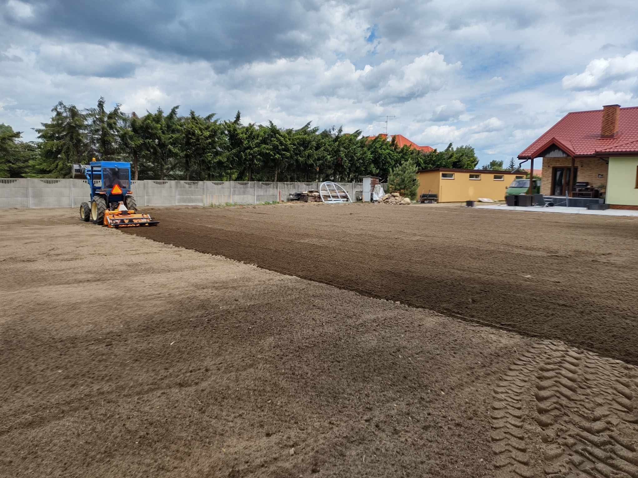Niebieski traktor z glebogryzarką przygotowuje ziemię pod trawnik na działce z domem i ogrodzeniem. Świeżo przekopana ziemia, ślady opon.