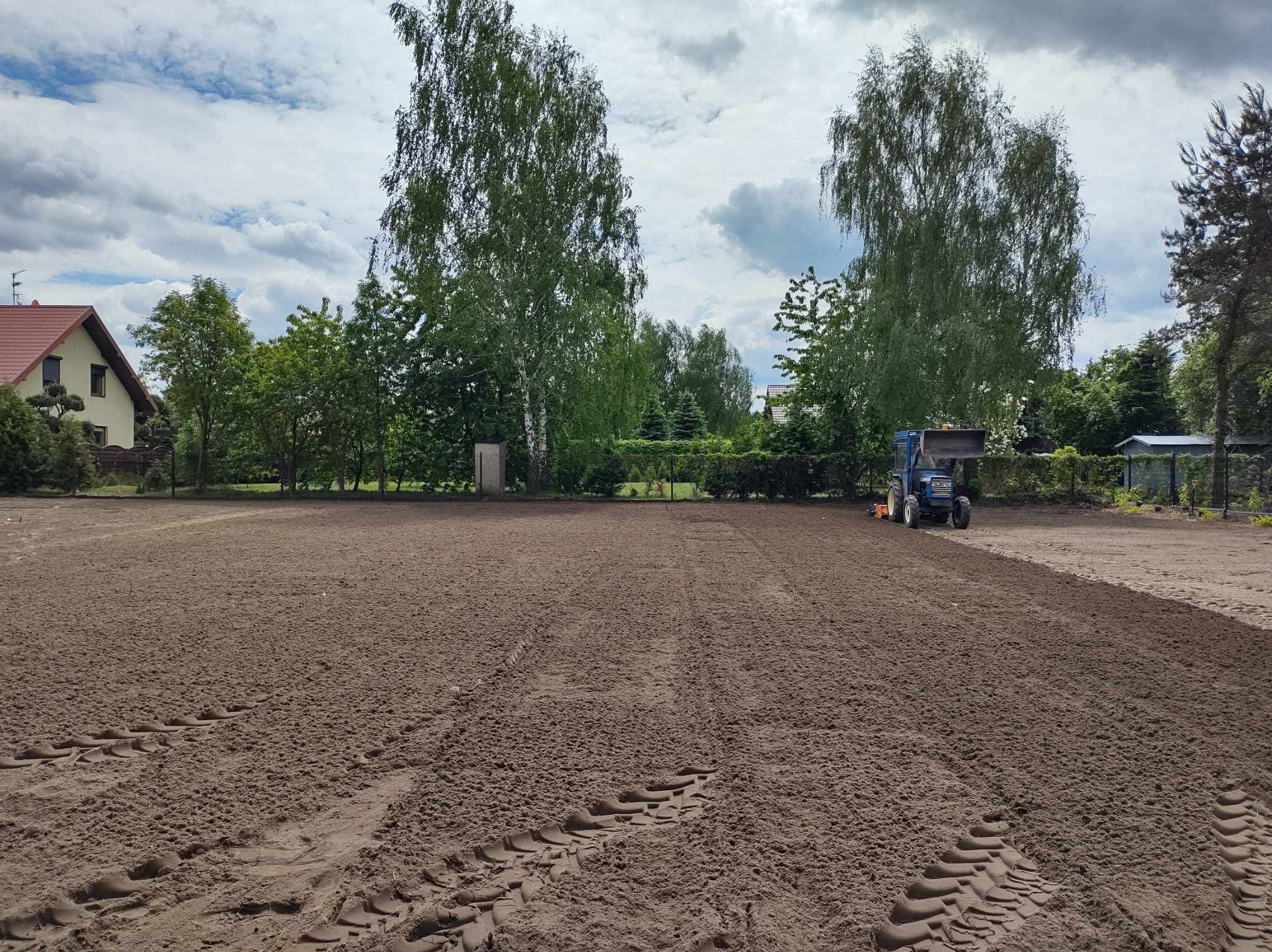 Przygotowanie terenu pod trawnik: wyrównana ziemia z widocznymi śladami opon traktora, drzewa w tle i budynek mieszkalny w oddali. Błękitne niebo z chmurami.