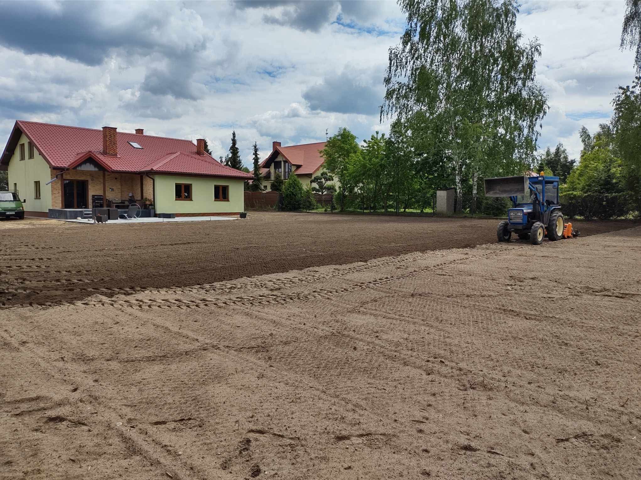 Przygotowanie gruntu pod trawnik: wyrównana ziemia z widocznymi śladami traktora na tle domu z czerwoną dachówką i zieleni drzew. Prace ziemne w toku.