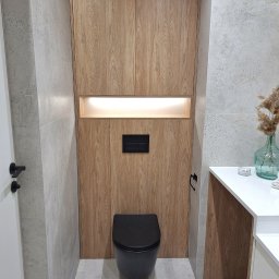 Ventures Michał Preś - Nowoczesna toaleta z zabudową meblową z drewnopodobną okleiną, podświetlana wnęka nad kompaktem WC, szara płytka podłogowa i ścienna, minimalistyczna armatura w kolorze czarnym.