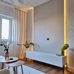 Ventures Michał Preś - Nowoczesny salon z białą szafką RTV, podświetleniem LED w ścianie, drewnianą podłogą, okrągłymi stolikami kawowymi i designerską lampą sufitową.