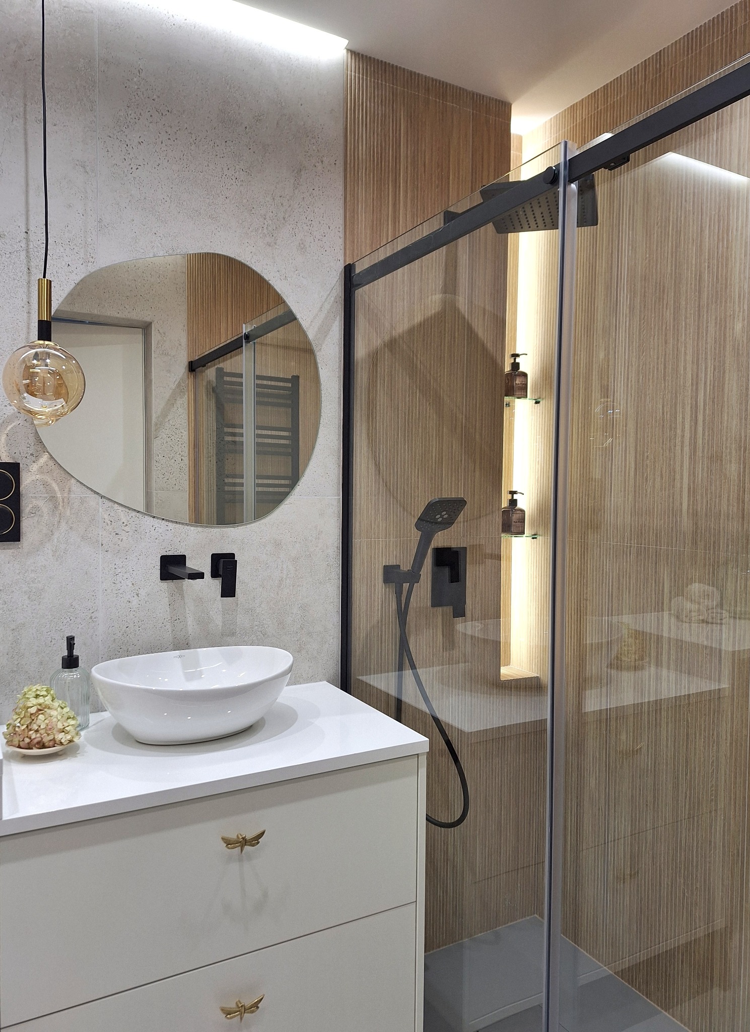 Nowoczesna łazienka z okrągłym lustrem, białą umywalką na szafce z ozdobnymi uchwytami w kształcie ważek i kabiną prysznicową z czarnymi detalami.