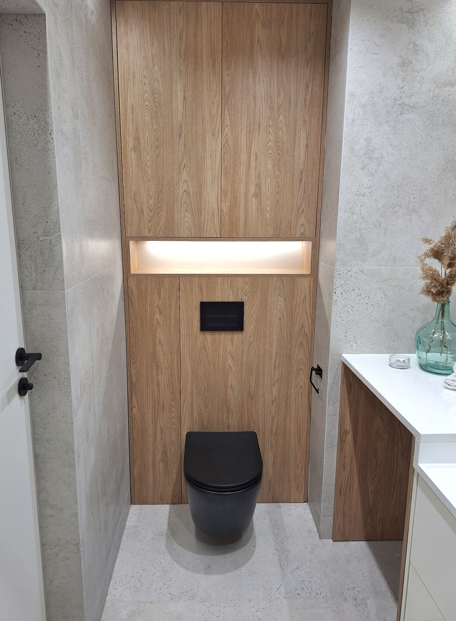Nowoczesna toaleta z zabudową meblową z drewnopodobną okleiną, podświetlana wnęka nad kompaktem WC, szara płytka podłogowa i ścienna, minimalistyczna armatura w kolorze czarnym.