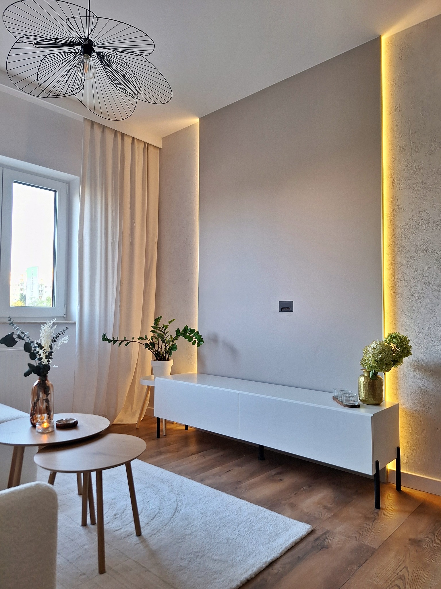 Nowoczesny salon z białą szafką RTV, podświetleniem LED w ścianie, drewnianą podłogą, okrągłymi stolikami kawowymi i designerską lampą sufitową.