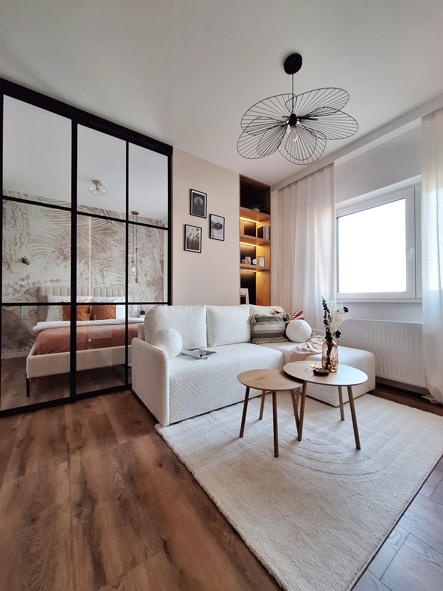 Jasny salon z drewnianą podłogą i białym dywanem, sofa z tkaniny boucle, stoliki kawowe, okno z białymi zasłonami, przeszklona ściana oddzielająca sypialnię z tapetą w motywy roślinne i łóżkiem...