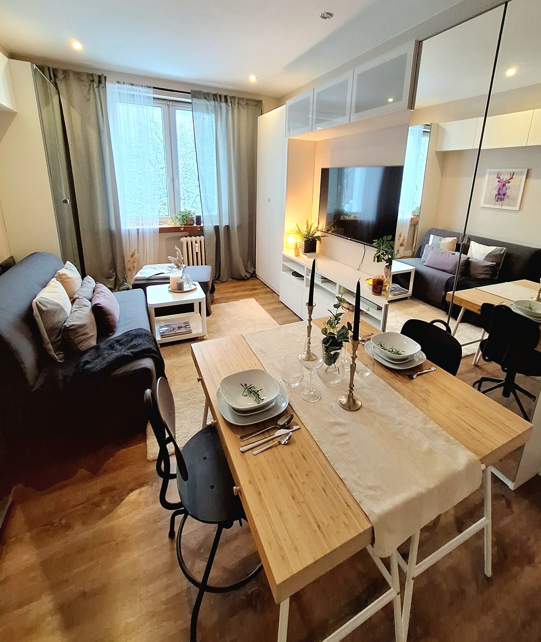 Ustawiony stół z drewnianym blatem, białym bieżnikiem, zastawą i świecznikami w jasnym, nowocześnie urządzonym salonie z szarą sofą i dużym oknem.