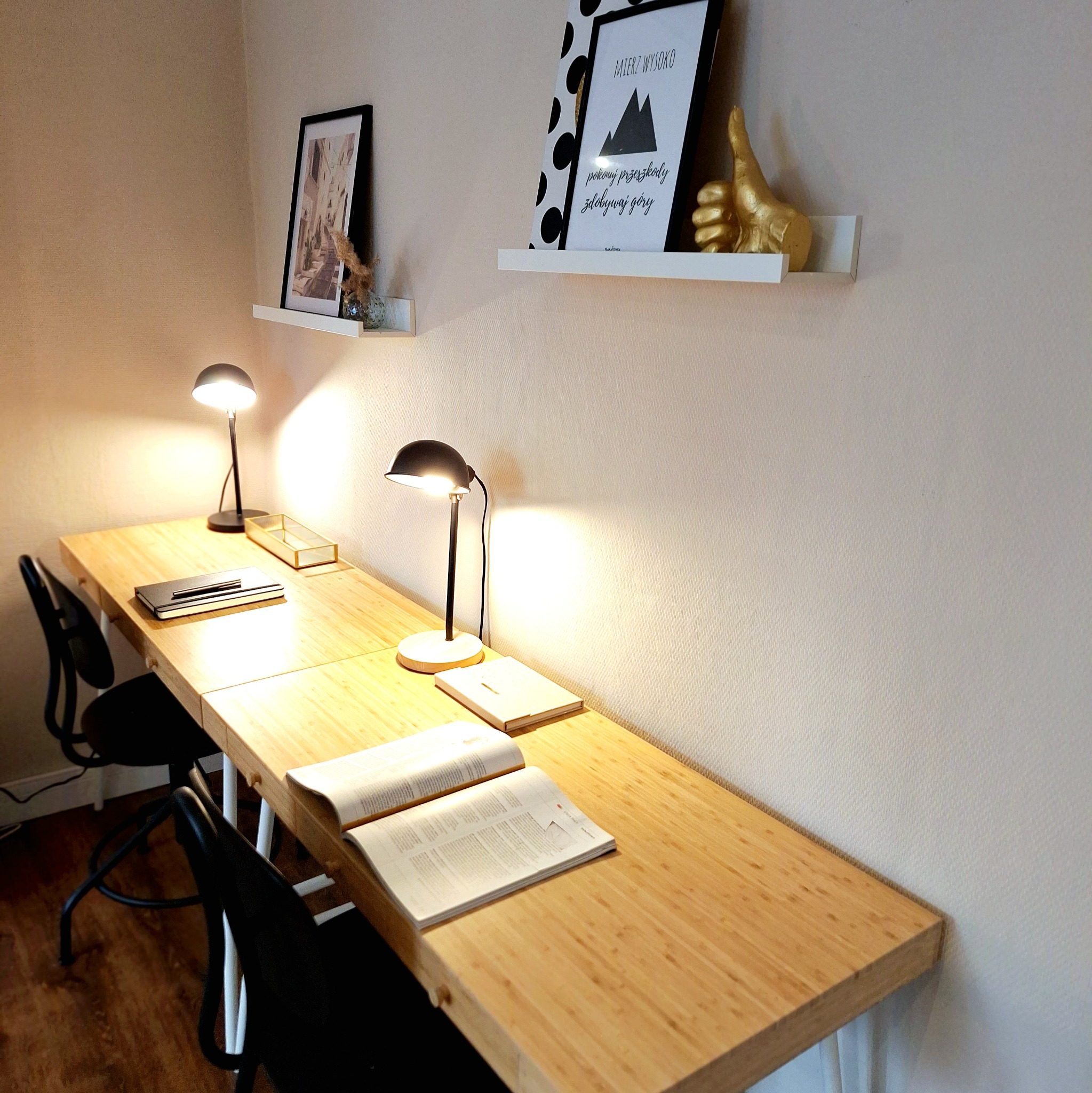 Długi, drewniany blat biurka z dwoma czarnymi krzesłami, oświetlony dwiema lampkami; na ścianie wiszą półki z dekoracjami, w tym złota figurka kciuka w górę.
