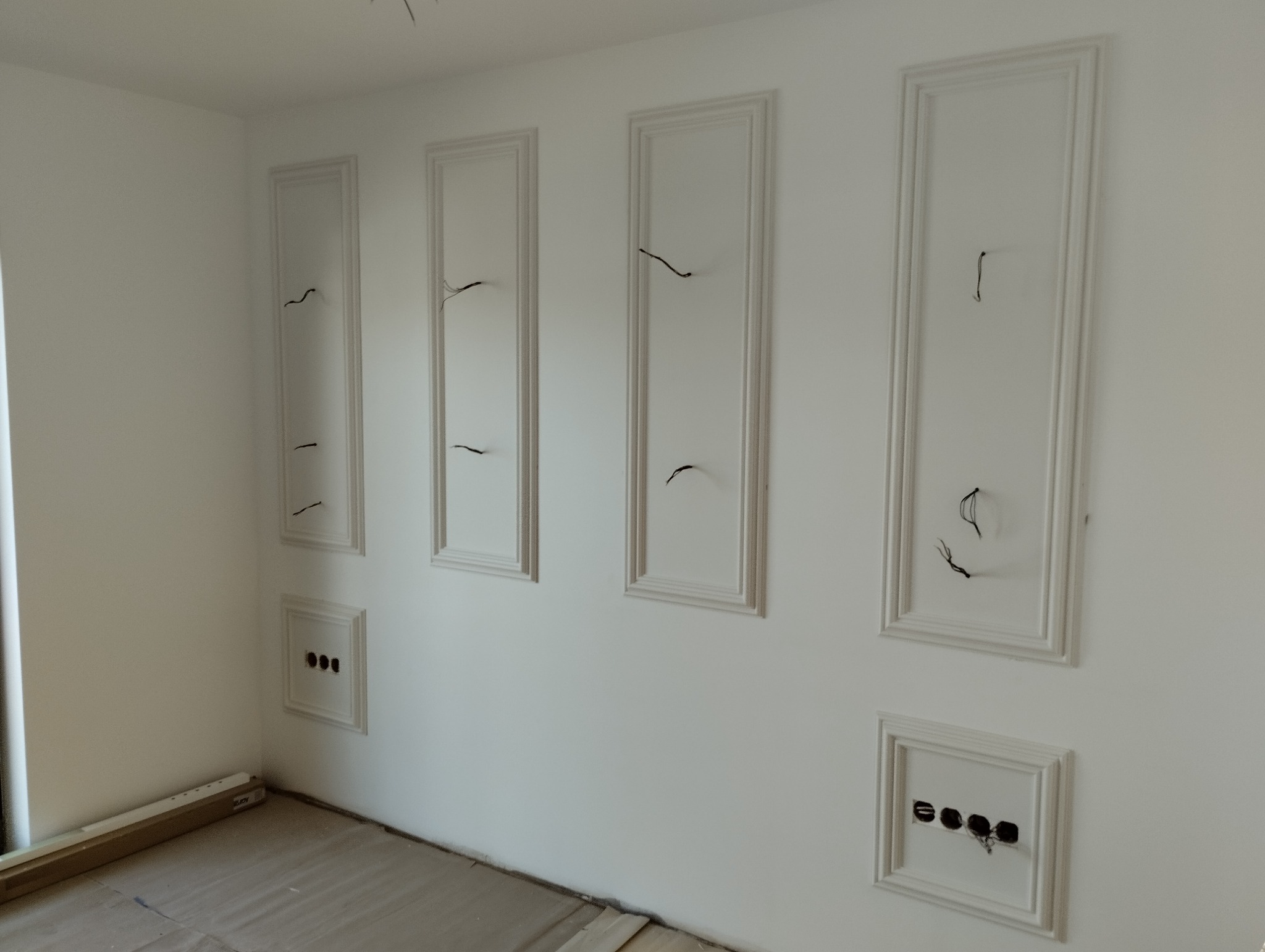 Białe ściany z ozdobnymi listwami i wyprowadzonymi przewodami elektrycznymi, przygotowane do montażu oświetlenia lub innych instalacji, widoczne gniazdka elektryczne bez osprzętu.