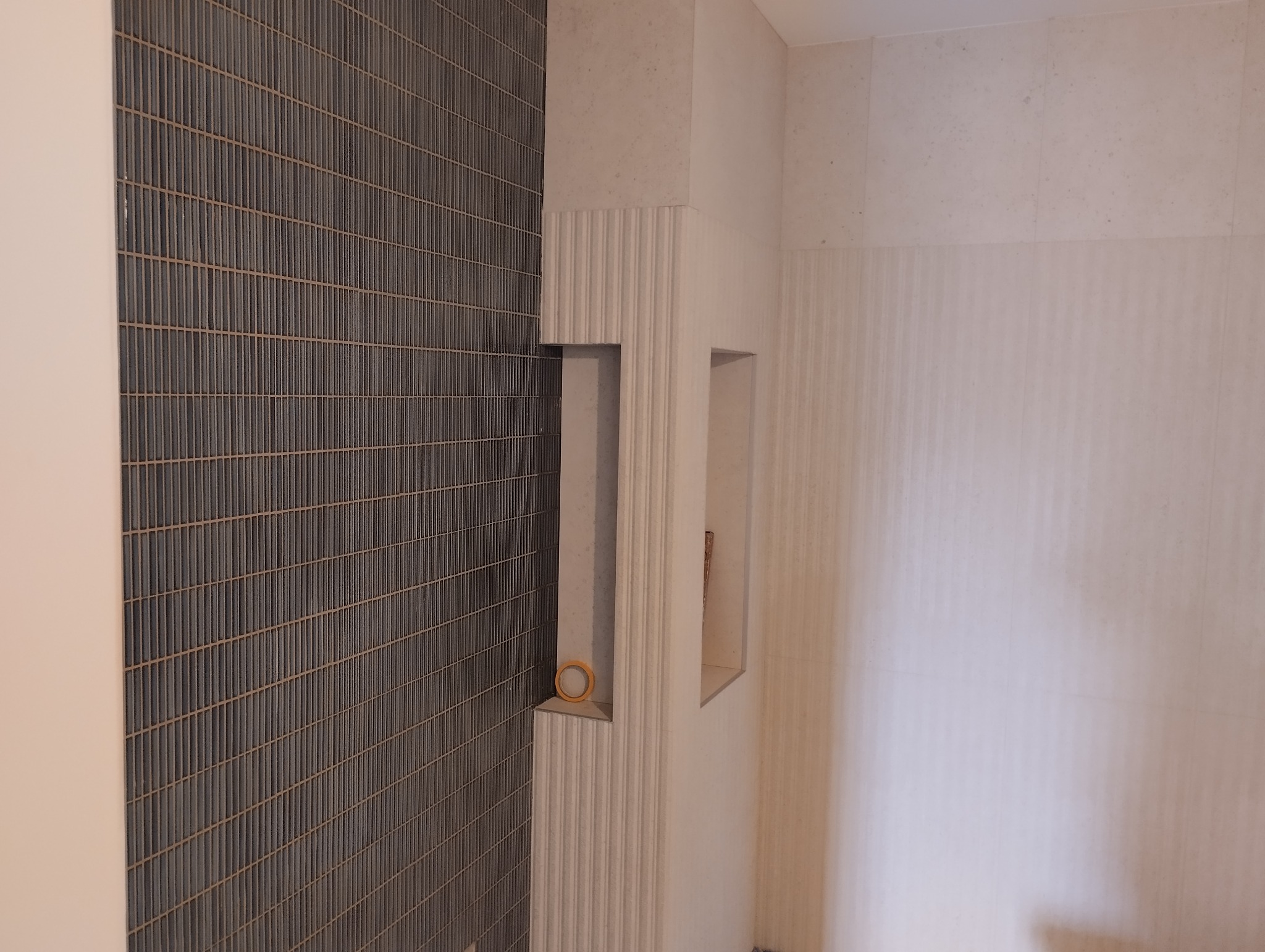 Ściana wyłożona wąskimi, ciemnymi płytkami, z wnękami wykończonymi jasnymi, pionowo żłobionymi panelami. Na jednej z półek leży rolka taśmy malarskiej.