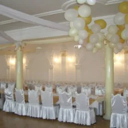 Uroczysta sala bankietowa z białymi krzesłami w pokrowcach, kolumnami z dekoracjami i balonami w kolorach białym i złotym pod sufitem.