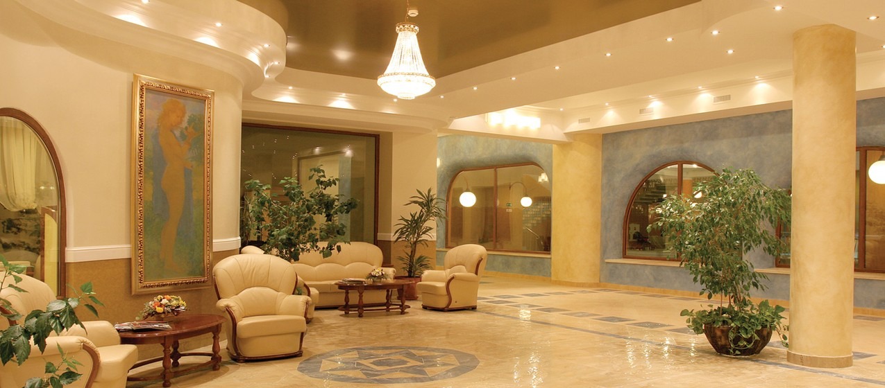 Eleganckie wnętrze holu z kremowymi meblami wypoczynkowymi, obrazem w złotej ramie, roślinami doniczkowymi i marmurową podłogą z dekoracyjnymi elementami, oświetlone kryształowym żyrandolem...