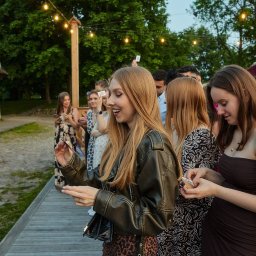 Iluzjonista Piotr Suder - Uśmiechnięta kobieta prezentuje trik z kartami podczas imprezy plenerowej, w tle obserwujący goście i dekoracyjne lampki.