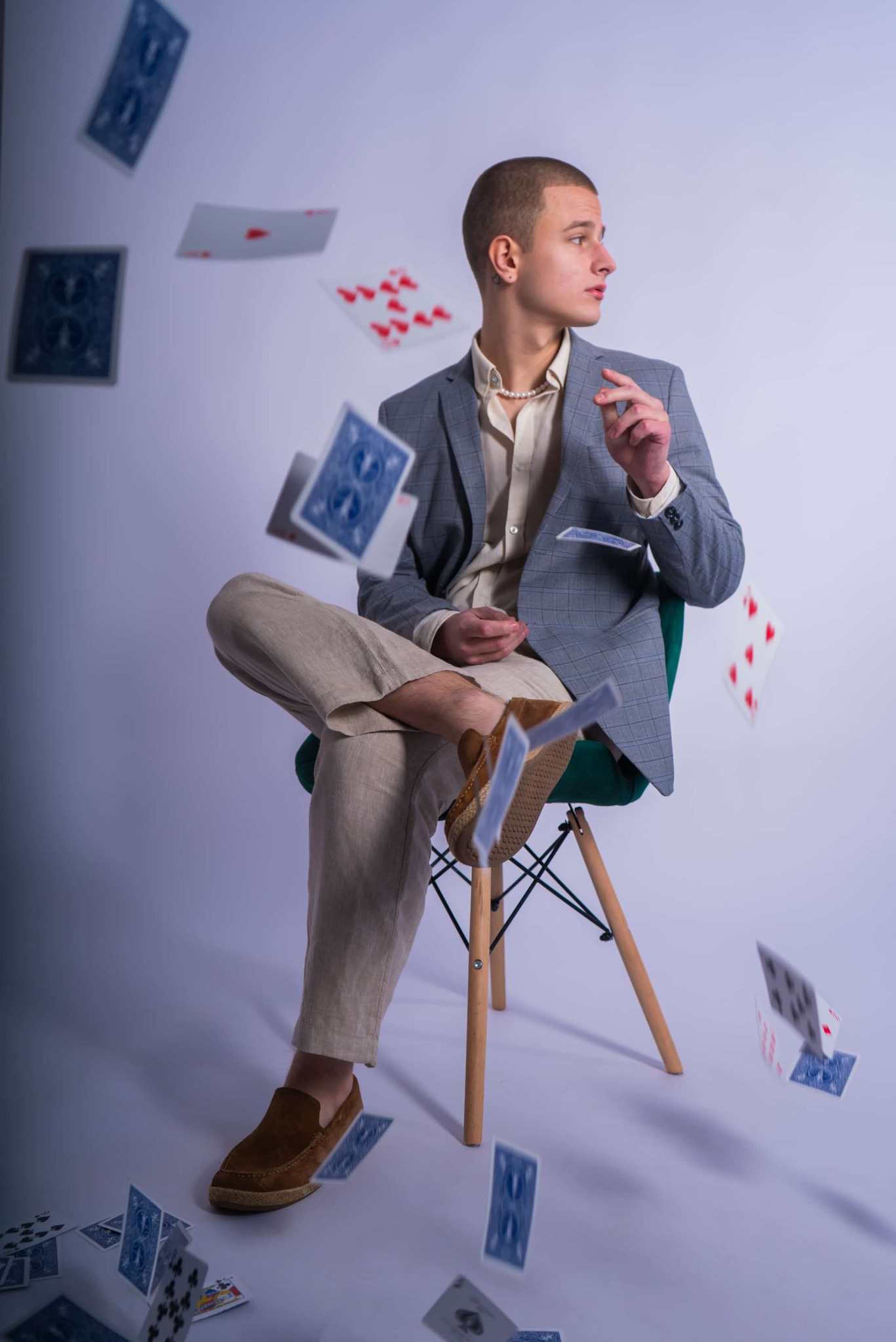 Młody mężczyzna w szarej marynarce siedzi na krześle, otoczony unoszącymi się kartami do gry, zamyślony wzrok skierowany w bok.