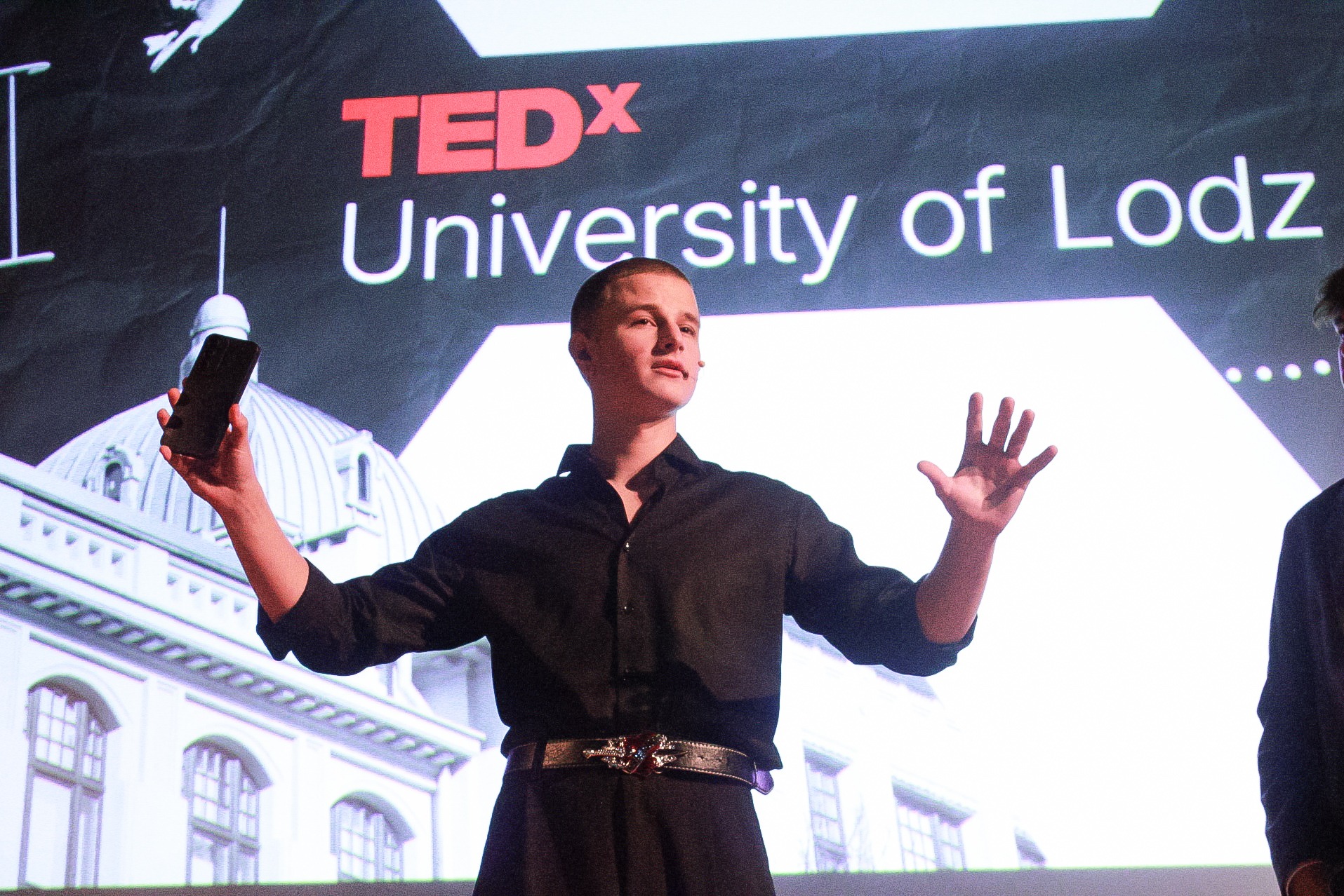 Prelegent w czarnej koszuli i ozdobnym pasku prezentuje na scenie z logiem TEDx University of Lodz, trzymając w jednej ręce telefon.