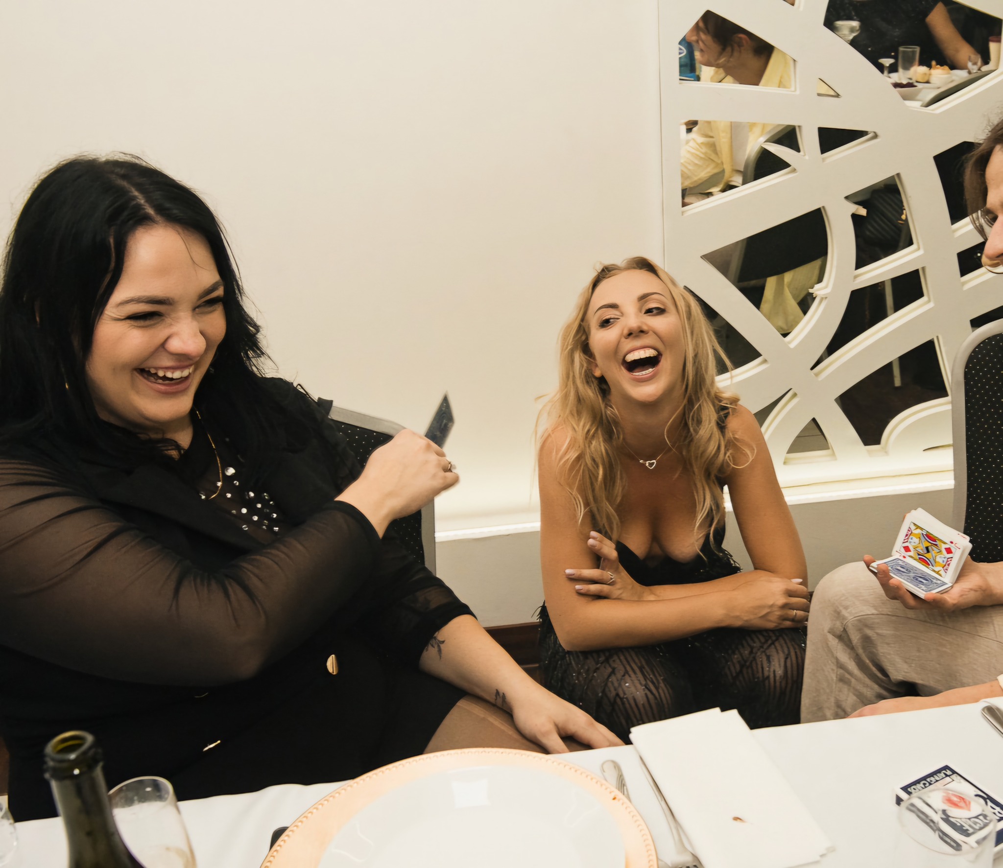 Iluzjonista prezentuje sztuczkę z kartami dwóm rozbawionym kobietom podczas przyjęcia; na stole leżą karty, talerze i butelka.