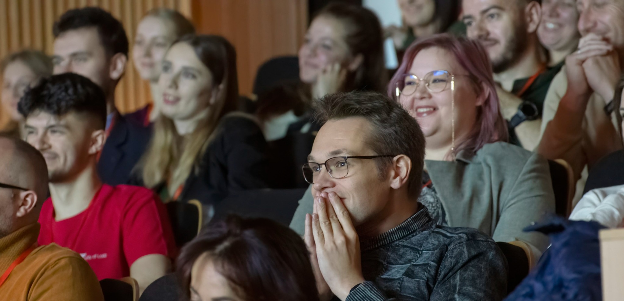 Widownia podczas występu, mężczyzna w okularach z rękami przy twarzy, w tle uśmiechnięte osoby.