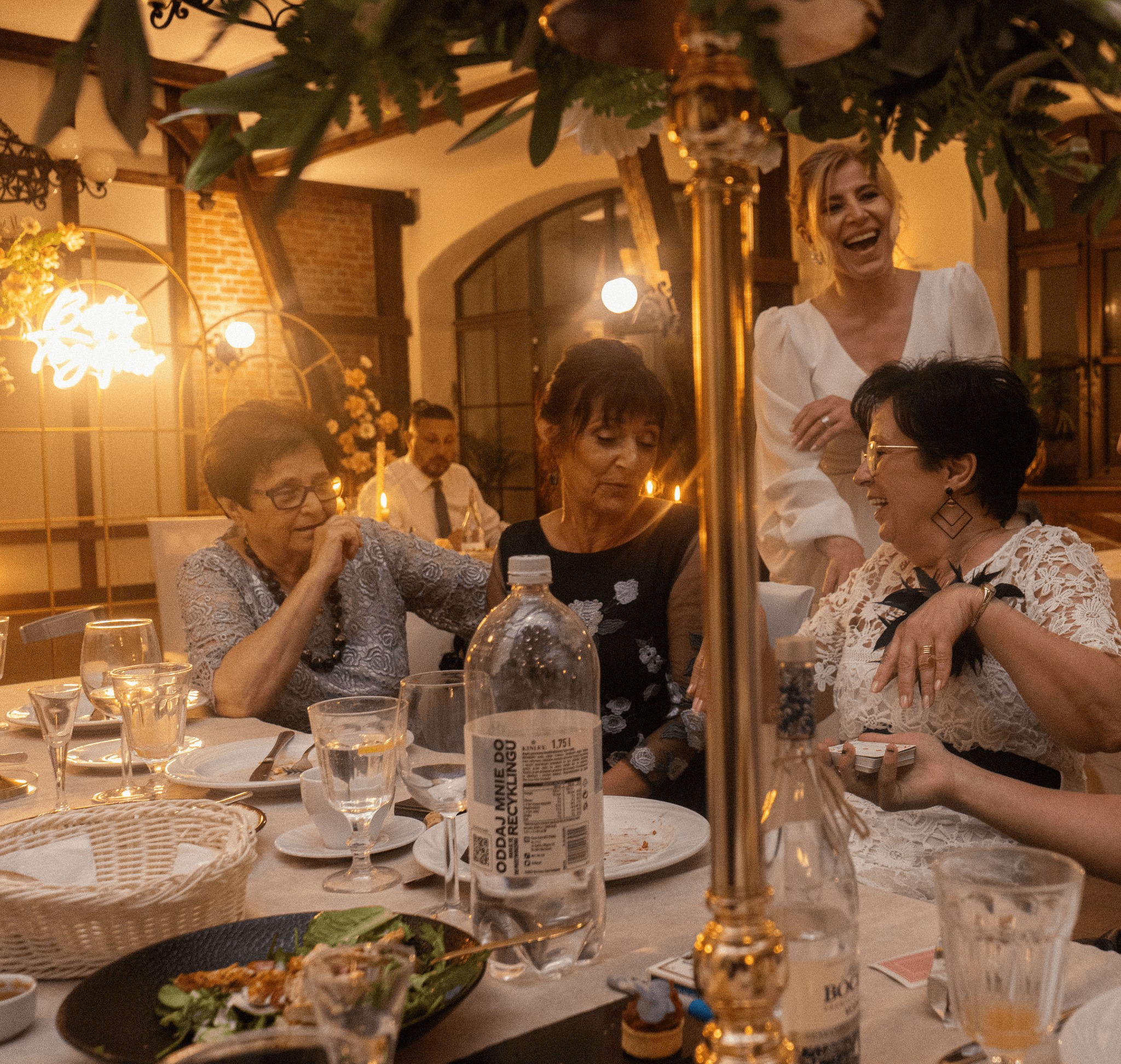 Goście weselni siedzą przy stole z dekoracjami kwiatowymi i złotymi świecznikami podczas przyjęcia, jedna z kobiet trzyma w rękach talię kart.