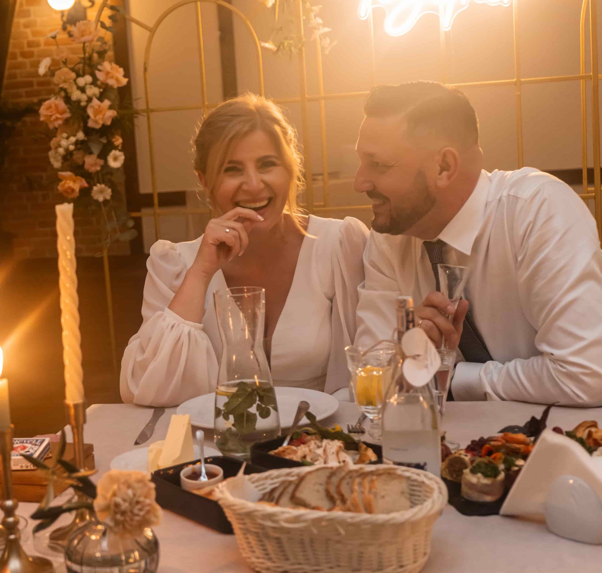 Para młoda siedząca przy stole podczas wesela, z dekoracjami kwiatowymi, świecami i potrawami, oświetlona ciepłym światłem.