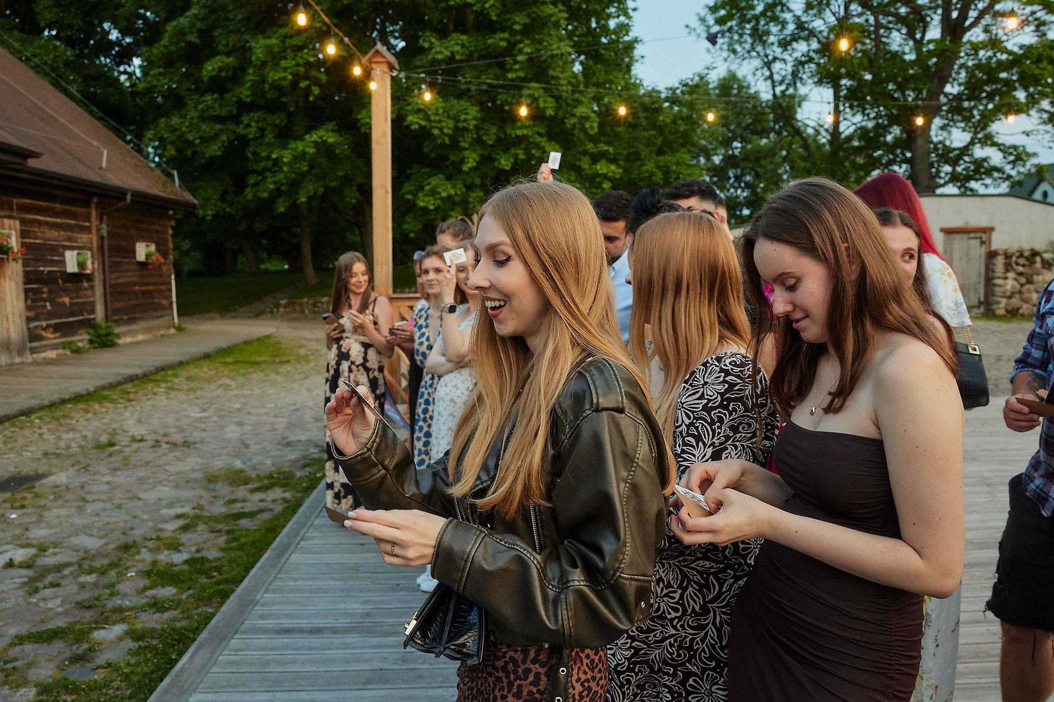 Uśmiechnięta kobieta prezentuje trik z kartami podczas imprezy plenerowej, w tle obserwujący goście i dekoracyjne lampki.