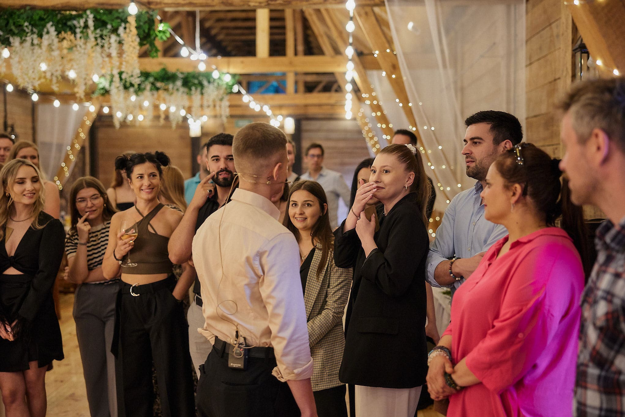 Uczestnicy wydarzenia w rustykalnej sali z dekoracją świetlną, mężczyzna z mikroportem tyłem, skupiona uwaga grupy, ciepłe oświetlenie.