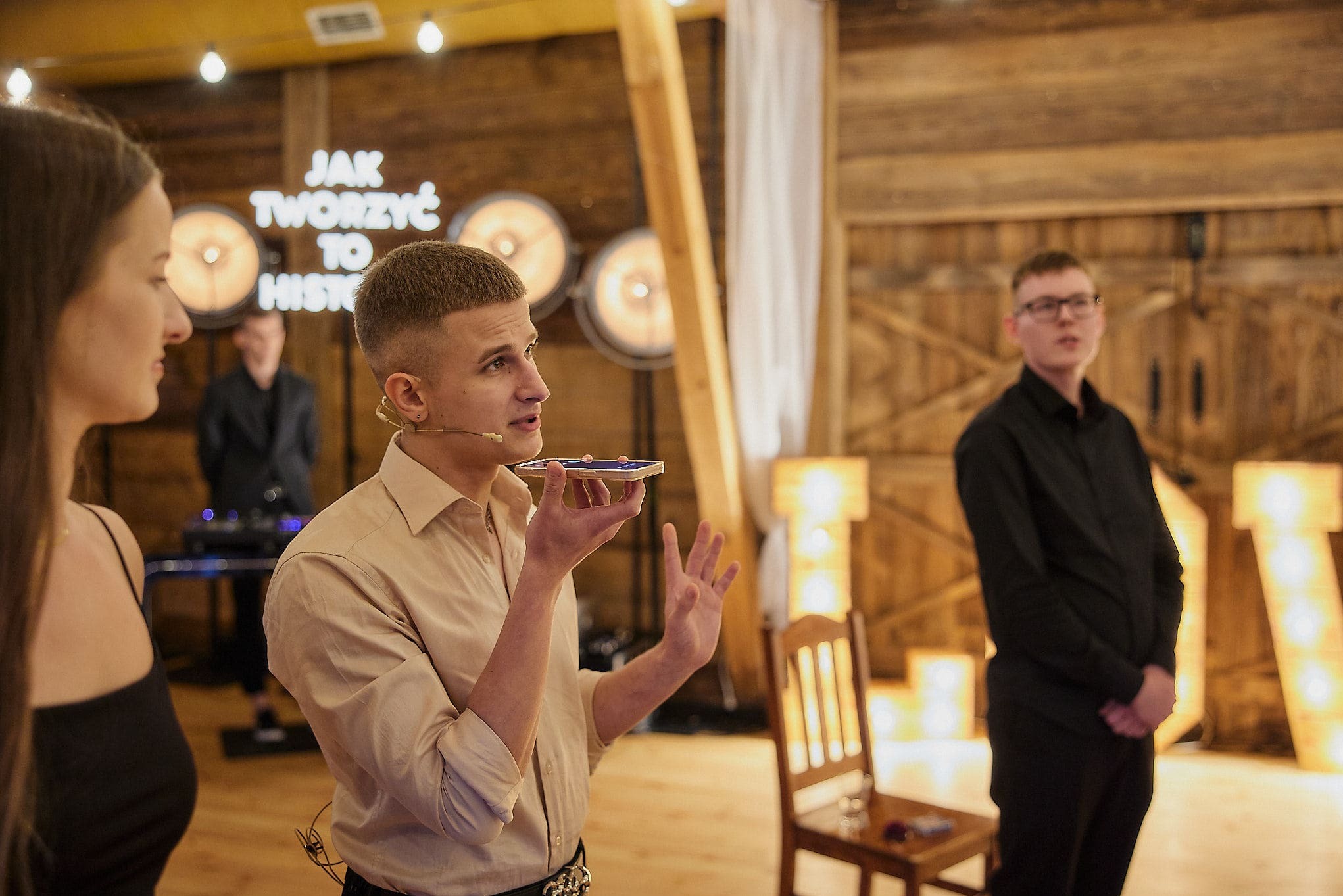 Młody mężczyzna z mikroportem gestykuluje, trzymając telefon przy ustach. W tle drewniana ściana z dekoracyjnym napisem i oświetleniem. Część występu podczas eventu.