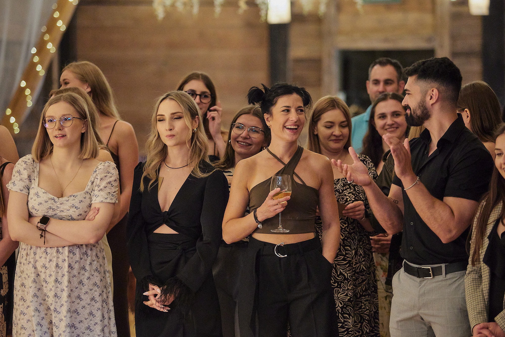 Uśmiechnięci goście na imprezie, kobieta z kieliszkiem w centrum, mężczyzna klaszcze, dekoracyjne oświetlenie w tle, eleganckie stroje.