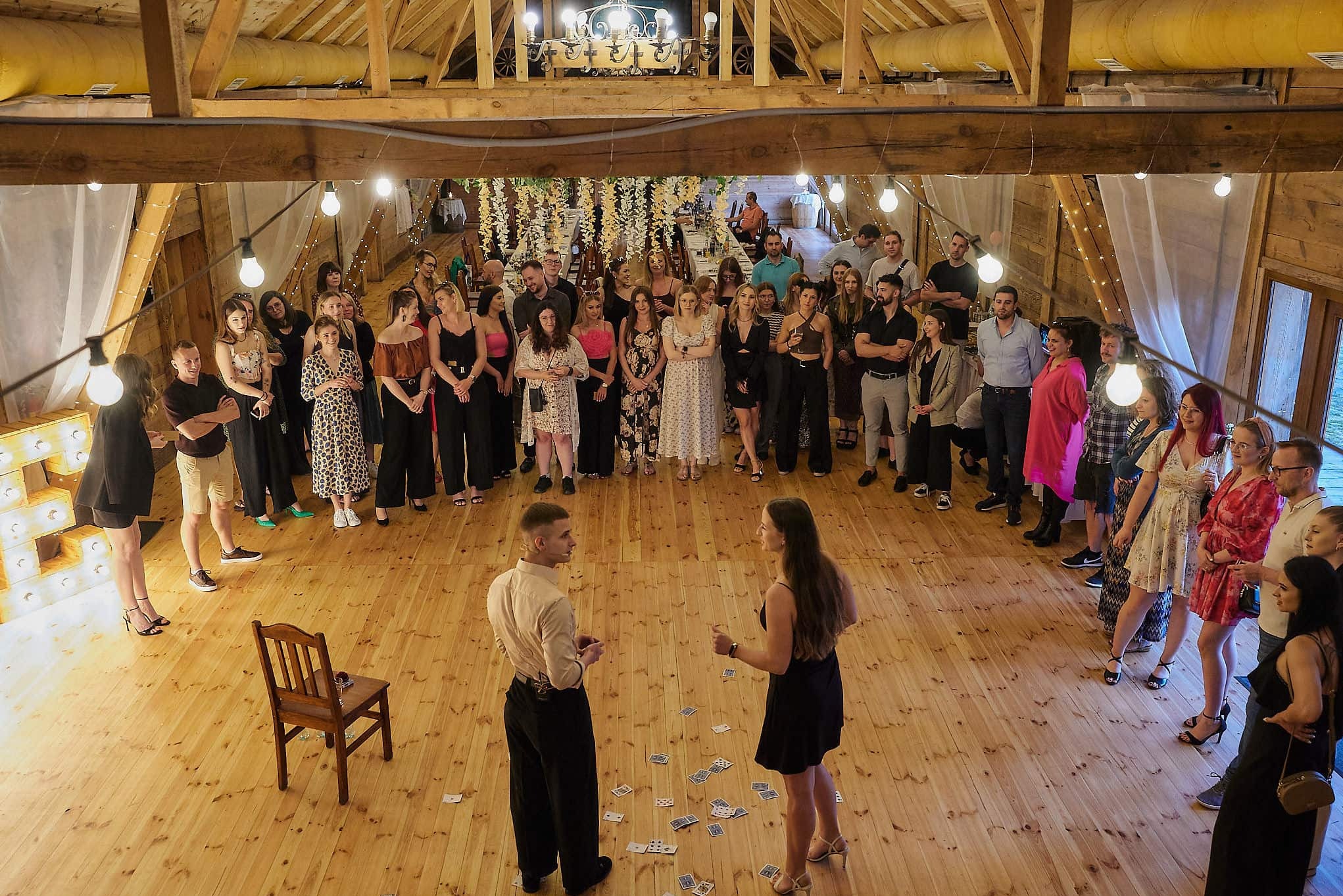 Iluzjonista prezentuje trik z kartami przed zgromadzoną publicznością w rustykalnej sali z drewnianą podłogą i dekoracjami świetlnymi.