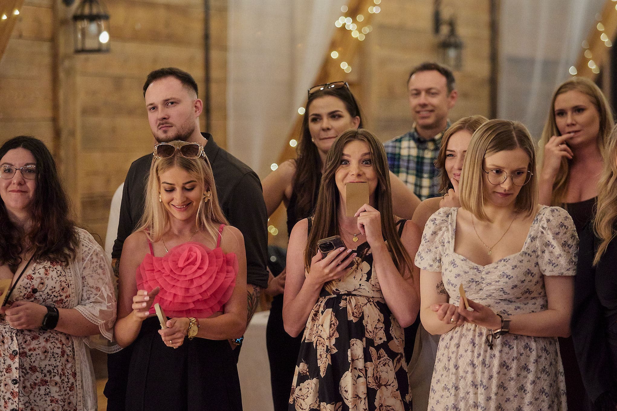 Grupa gości na evencie w sali z drewnianymi ścianami i dekoracyjnymi światłami. Kobieta w kwiecistej sukience trzyma kartkę przy twarzy.