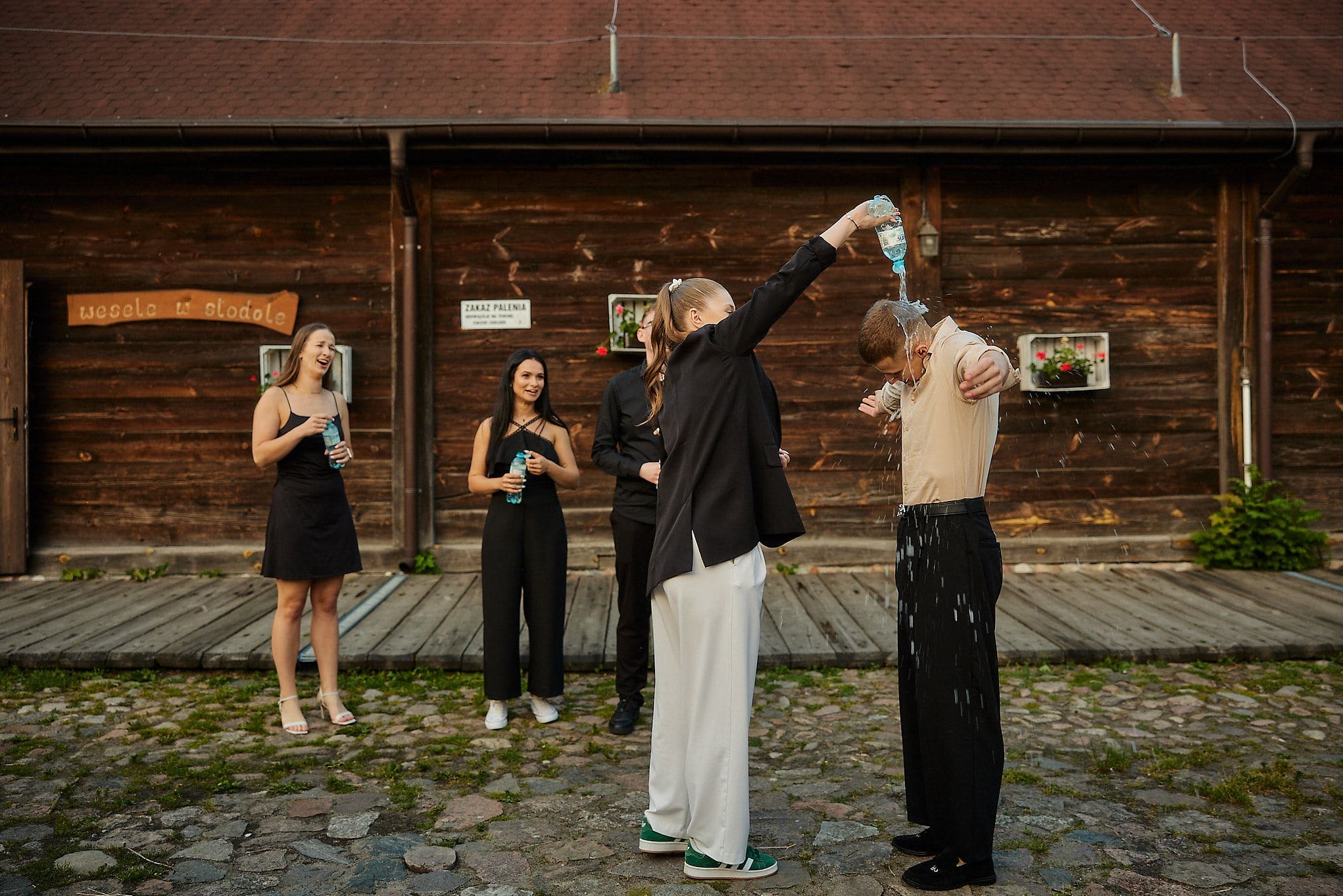 Zabawna scena z imprezy plenerowej: kobieta oblewa mężczyznę wodą z butelki na tle drewnianego budynku z napisem 'Wesele w stodole'.