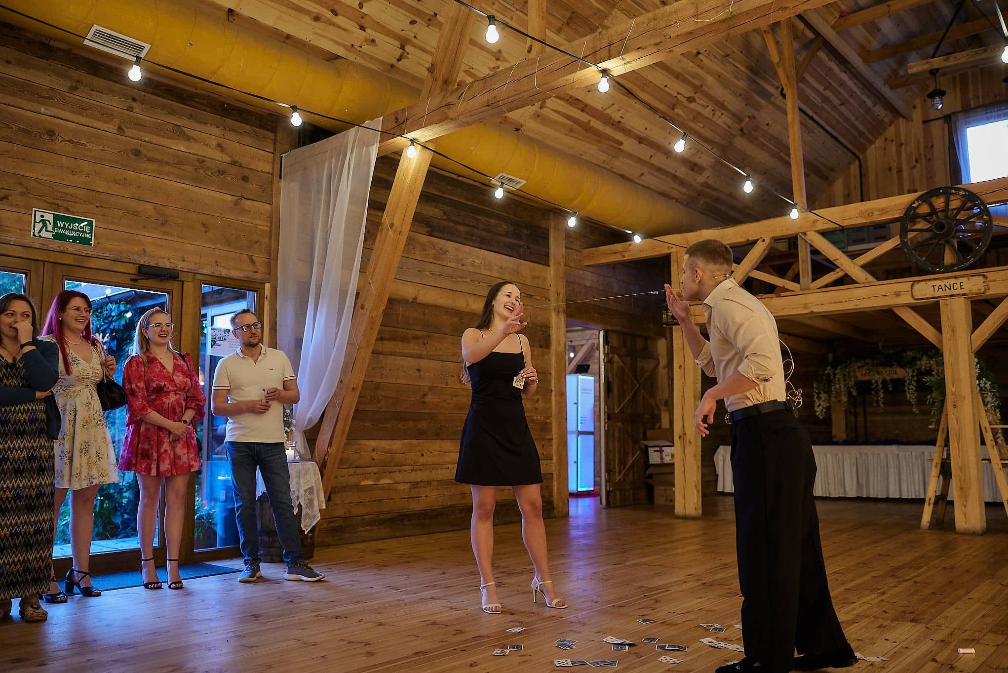 Iluzjonista prezentuje trik z kartami na drewnianym parkiecie, publiczność obserwuje z zaciekawieniem. Dekoracje rustykalne, ciepłe oświetlenie.