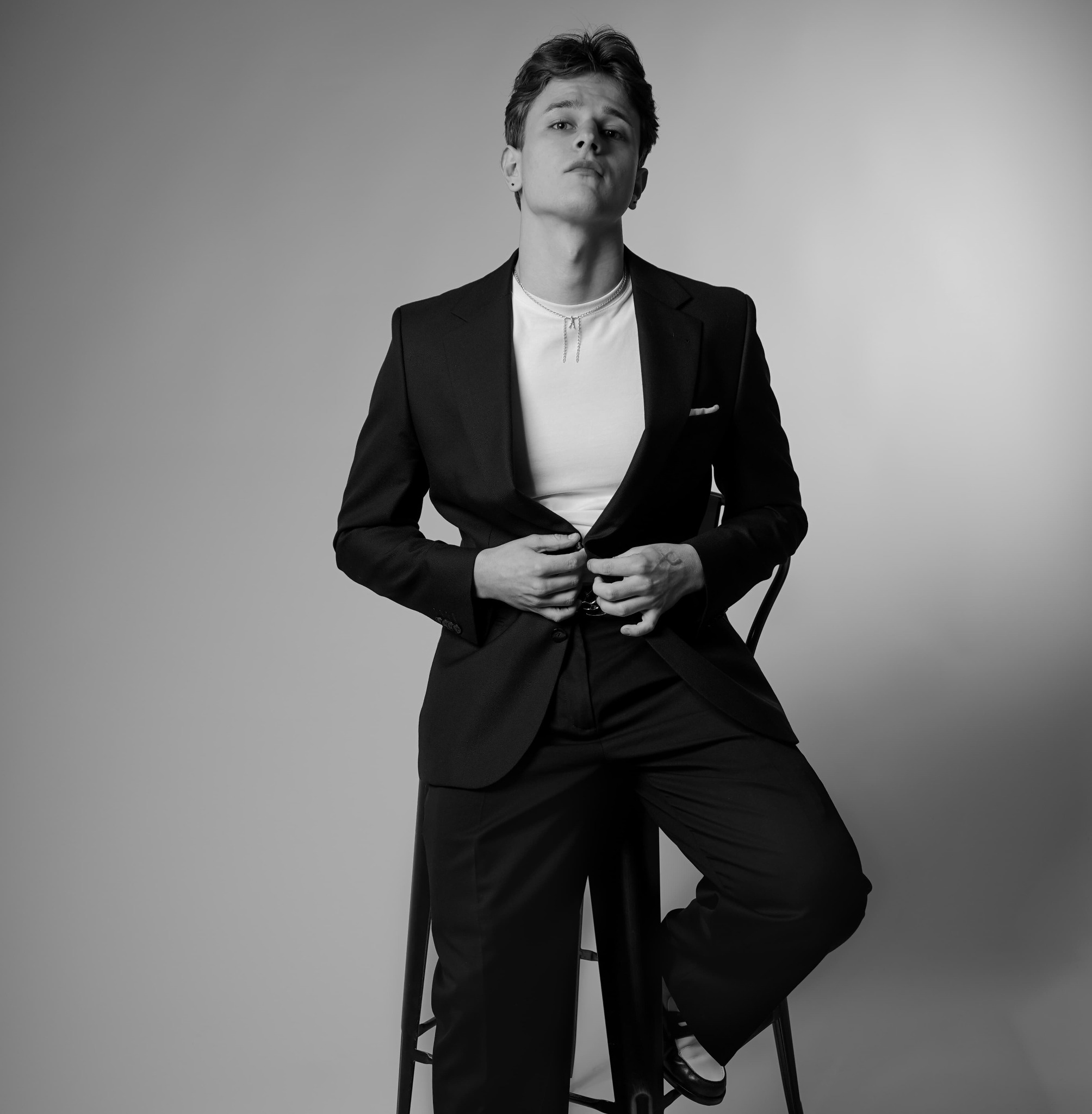 Czarno-białe zdjęcie młodego mężczyzny w garniturze, siedzącego na krześle i zapinającego marynarkę, z poważnym wyrazem twarzy i subtelnym oświetleniem.