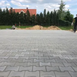 Ułożona szara kostka brukowa typu 'kość' na posesji, widoczny pracownik w trakcie pracy, w tle pryzma piasku, taczka i dom z czerwoną dachówką.