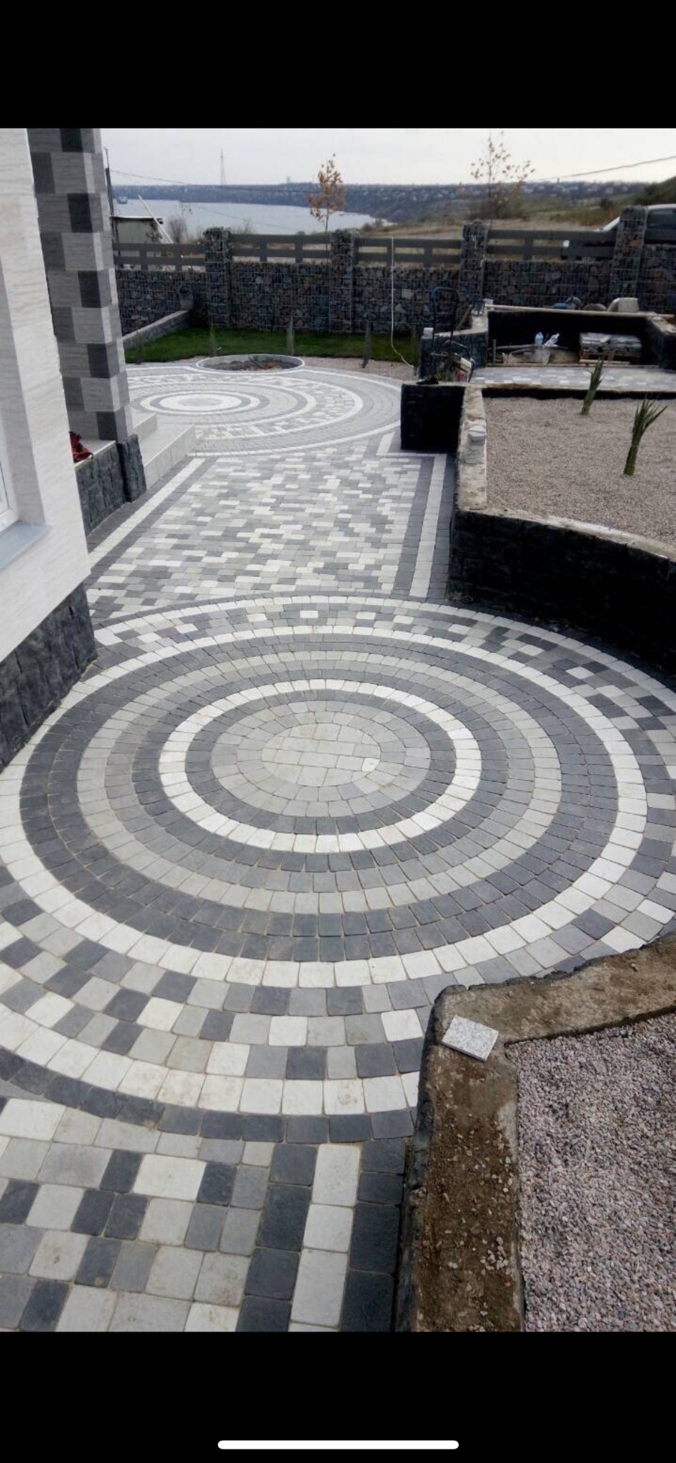 Ogród z dekoracyjną nawierzchnią z kostki brukowej ułożonej w koncentryczne okręgi i kwadraty, widok na jezioro w tle.
