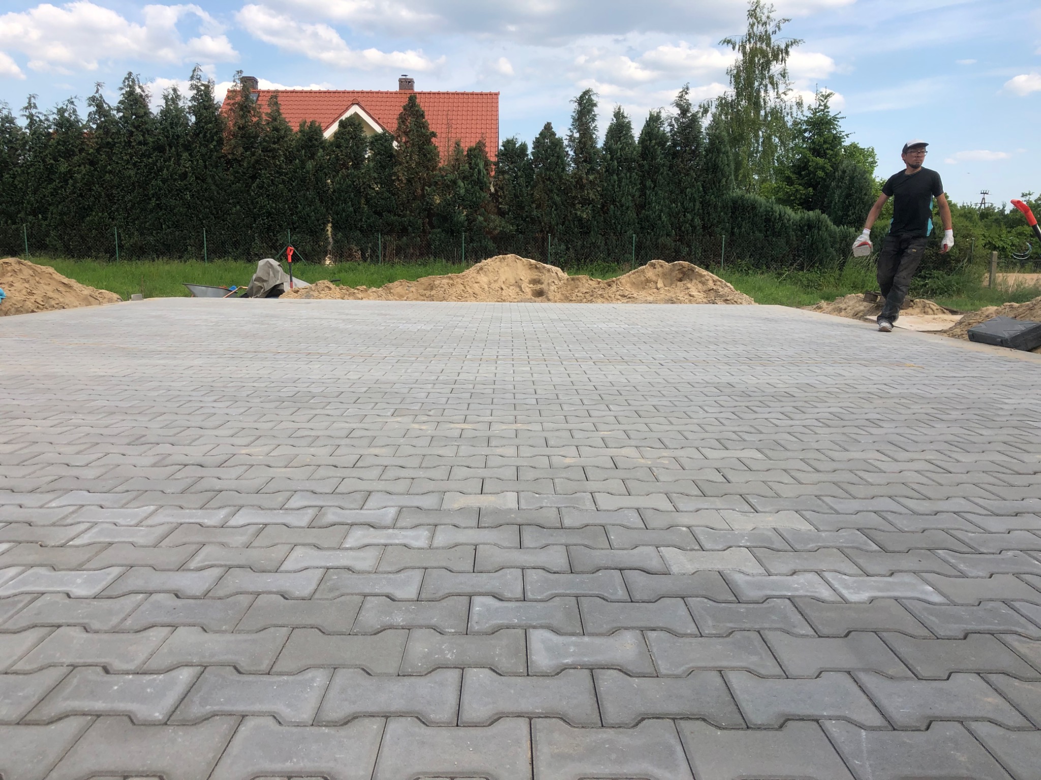 Ułożona szara kostka brukowa typu 'kość' na posesji, widoczny pracownik w trakcie pracy, w tle pryzma piasku, taczka i dom z czerwoną dachówką.