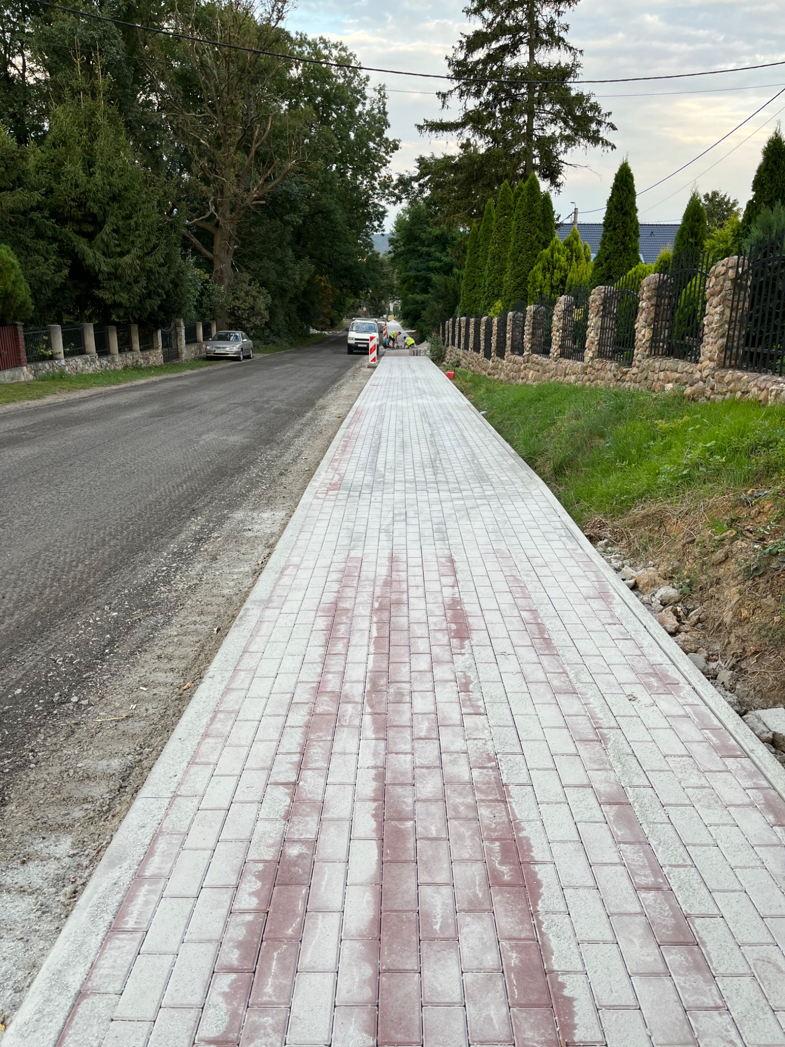 Długa, nowo ułożona ścieżka z kostki brukowej wzdłuż drogi, z widocznymi śladami wilgoci i dwoma pracownikami w oddali, otoczona zielenią i ogrodzeniem z kamienia.