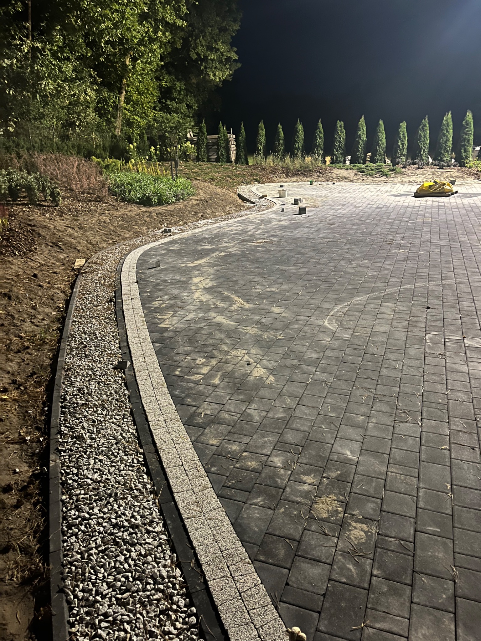 Ciemnoszare kostki brukowe ułożone na posesji, obrzeża wykończone jasną kostką i szarym żwirem, w tle rząd tuj i oświetlenie nocne.