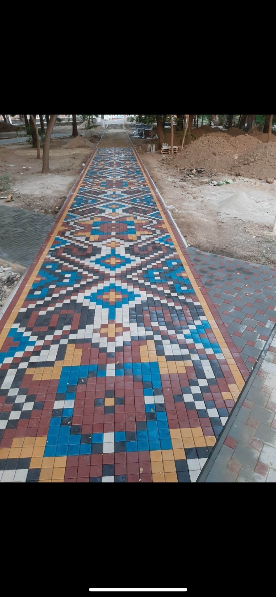 Długa ścieżka z kostki brukowej ułożonej we wzór geometryczny z użyciem kolorów: czerwonego, niebieskiego, żółtego, białego i czarnego, prowadząca przez teren zielony z drzewami i usypaną ziemią.