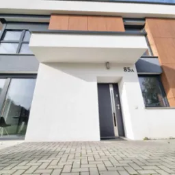 Nowoczesny dom jednorodzinny z elewacją w odcieniach bieli, szarości i drewna, z numerem 85a nad wejściem i brukowanym podjazdem.