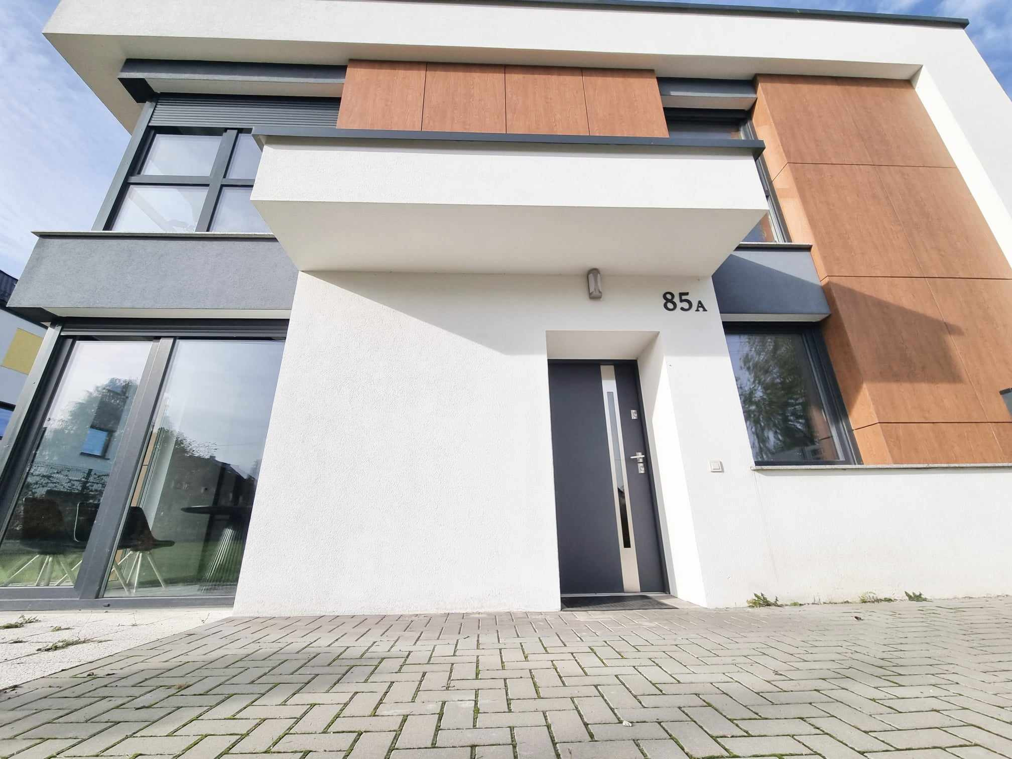 Nowoczesny dom jednorodzinny z elewacją w odcieniach bieli, szarości i drewna, z numerem 85a nad wejściem i brukowanym podjazdem.