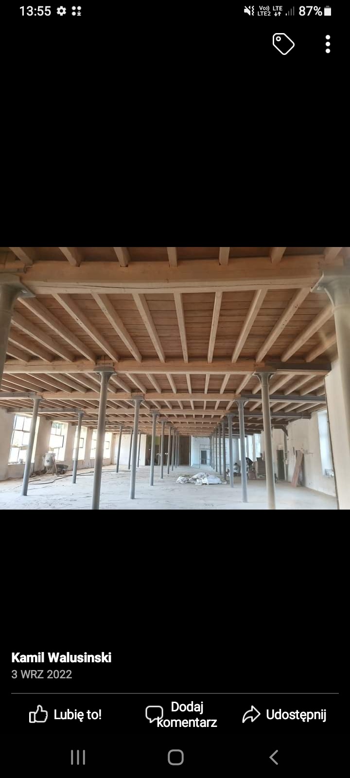 Wnętrze hali przemysłowej z drewnianym stropem podpartym rzędami metalowych kolumn, widoczne okna i resztki materiałów budowlanych na podłodze.