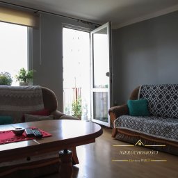 Biuro Nieruchomości Dariusz Wika - Jasne wnętrze salonu z balkonem, dwa brązowe fotele z poduszkami, drewniany stolik kawowy z pilotami, kwiaty w oknie, parkiet. Widok z mieszkania na zewnątrz.