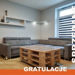 Biuro Nieruchomości Dariusz Wika - Jasne wnętrze salonu z szarą sofą narożną i stolikiem z palet. Na ścianie półki, okno z żaluzjami. Po prawej stronie napis 'Sprzedane' i 'Gratulacje'.