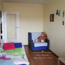 Pokój dziecięcy z rozkładaną sofą z kolorowymi wzorami, niebieskim fotelem z pluszowym misiem, dekoracjami na ścianie i drewnianą podłogą.